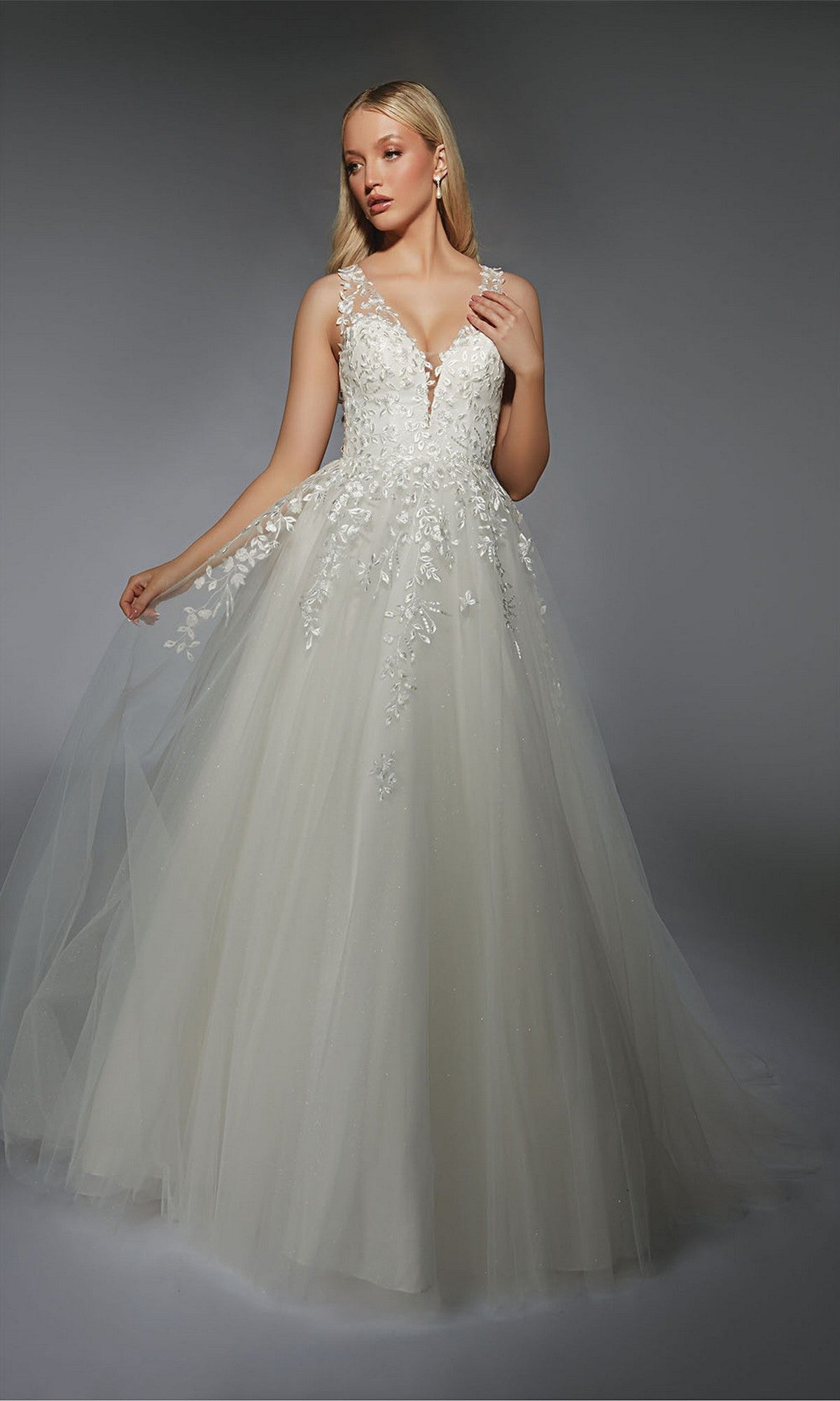 A woman models the Alyce 7128 Long White Dress, featuring delicate lace appliqué and a flowing tulle skirt, set against a plain gray background.